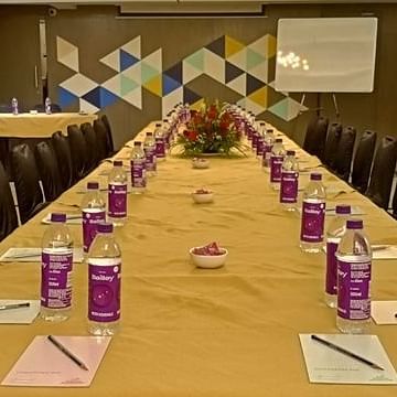 A long conference table set up for a meeting. There are water bottles, notepads, and pens arranged neatly at each seat. A whiteboard stands in the background, and the walls are decorated with a geometric pattern.